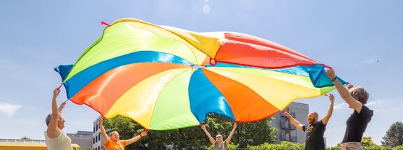 Eine Gruppe Rehabilitand*innen halten eine bunten "Fallschirm" durch Bewegung in der Luft. Die Sonne scheint. 