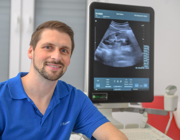 Dr. med. Michael Korten sitzt vor einem Ultraschallbild, welches am Computer angezeigt wird und lächelt in die Kamera. 