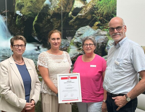 Die Klinikleitung der Gelderland-Klinik steht im Foyer der Reha-Klinik vor einer grünen Wand mit Wasserfall-Foto - von links zu sehen sind: Susanne Grohmann, Diplom-Psychologin, Dr. med. Sonja Höhndorf, Ärztliche Direktorin, die einen Rahmen mit der stern-Auszeichnung in ihren Händen hält, Karla Bergers, Direktorin für Pflege und Organisationsmanagement der Gelderland-Klinik und Ludger Broeckmann, Kaufmännischer Direktor des Gesundheitscampus St.-Clemens Geldern.