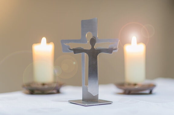 In der Mitte sieht man eine Person die die Arme ausbreitet aus Metall in einem kleinen Format und daneben stehen zwei Kerzen.