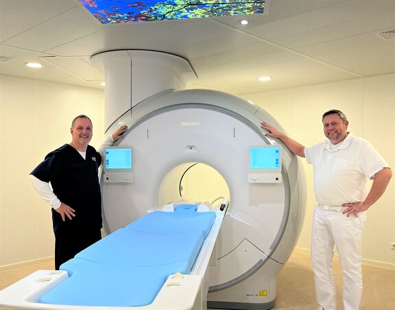 MTR Leiter und Chefarzt stehen rechts und links von einem MRT-Gerät und lächeln in die Kamera